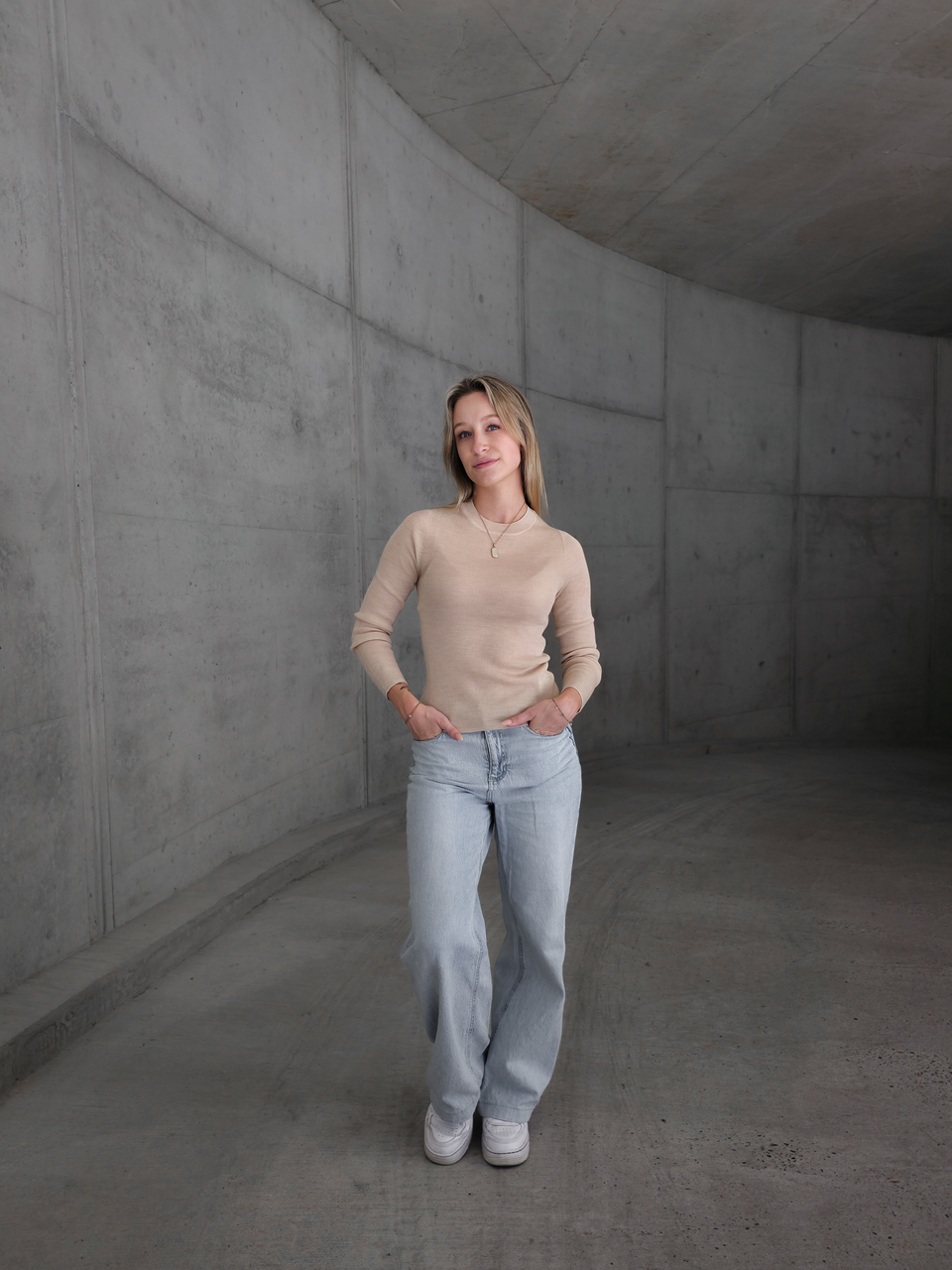 Danja im beigen Merino-Longsleeve in XS als Ganzkörperansicht vor Beton.