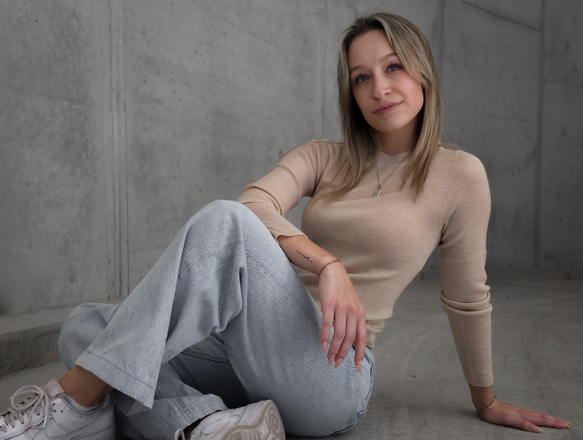 Danja sitzt im beigen Merino-Longsleeve in XS vor einer Betonwand.